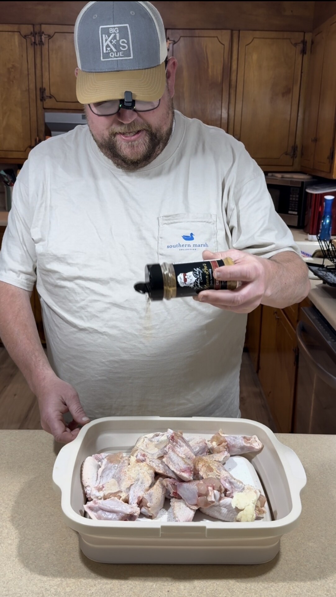 A man wearing a hat and glasses sprinkles seasoning from a bottle onto raw chicken pieces in a baking dish on a kitchen counter. Wooden cabinets and various kitchen items are visible in the background.