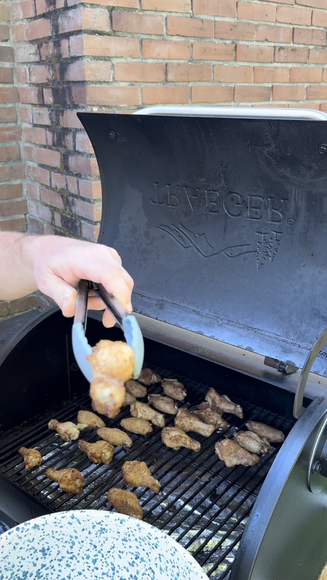 A hand holds metal tongs with a cooked chicken wing above a grill filled with several grilled chicken wings. The grill lid is open, revealing the brand name LIVECED. Brick wall is visible in the background.