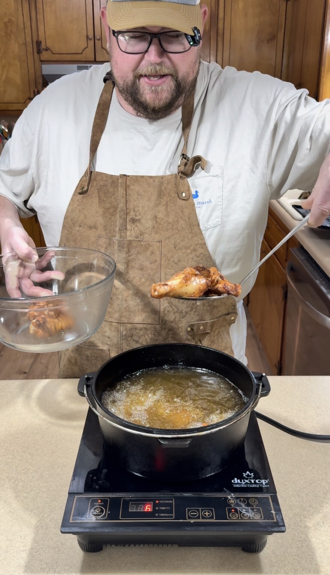 A man wearing a cap, glasses, and a brown apron lifts a piece of fried chicken from a pot of hot oil on an induction cooktop in a kitchen, holding a glass bowl in his other hand.