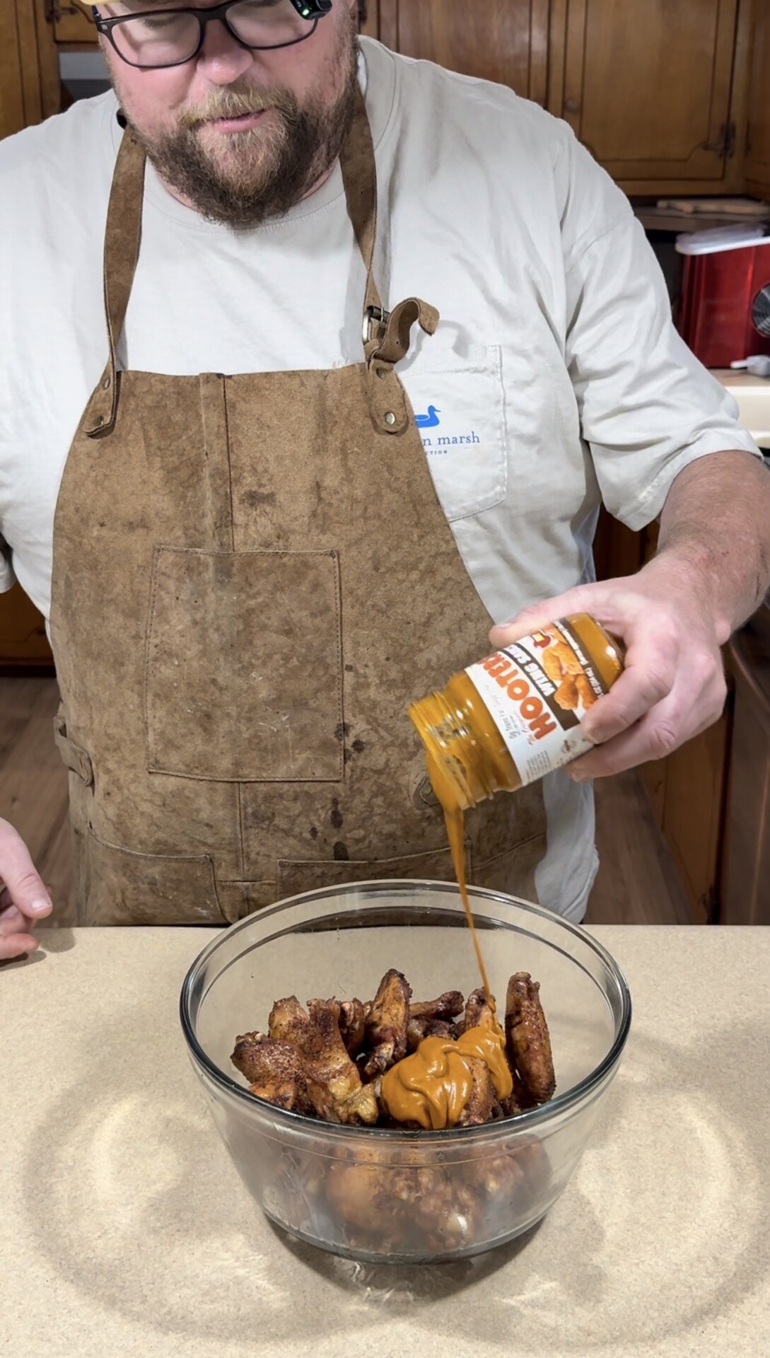 A person wearing a brown apron pours a jar of orange sauce over chicken wings in a glass bowl on a kitchen counter.