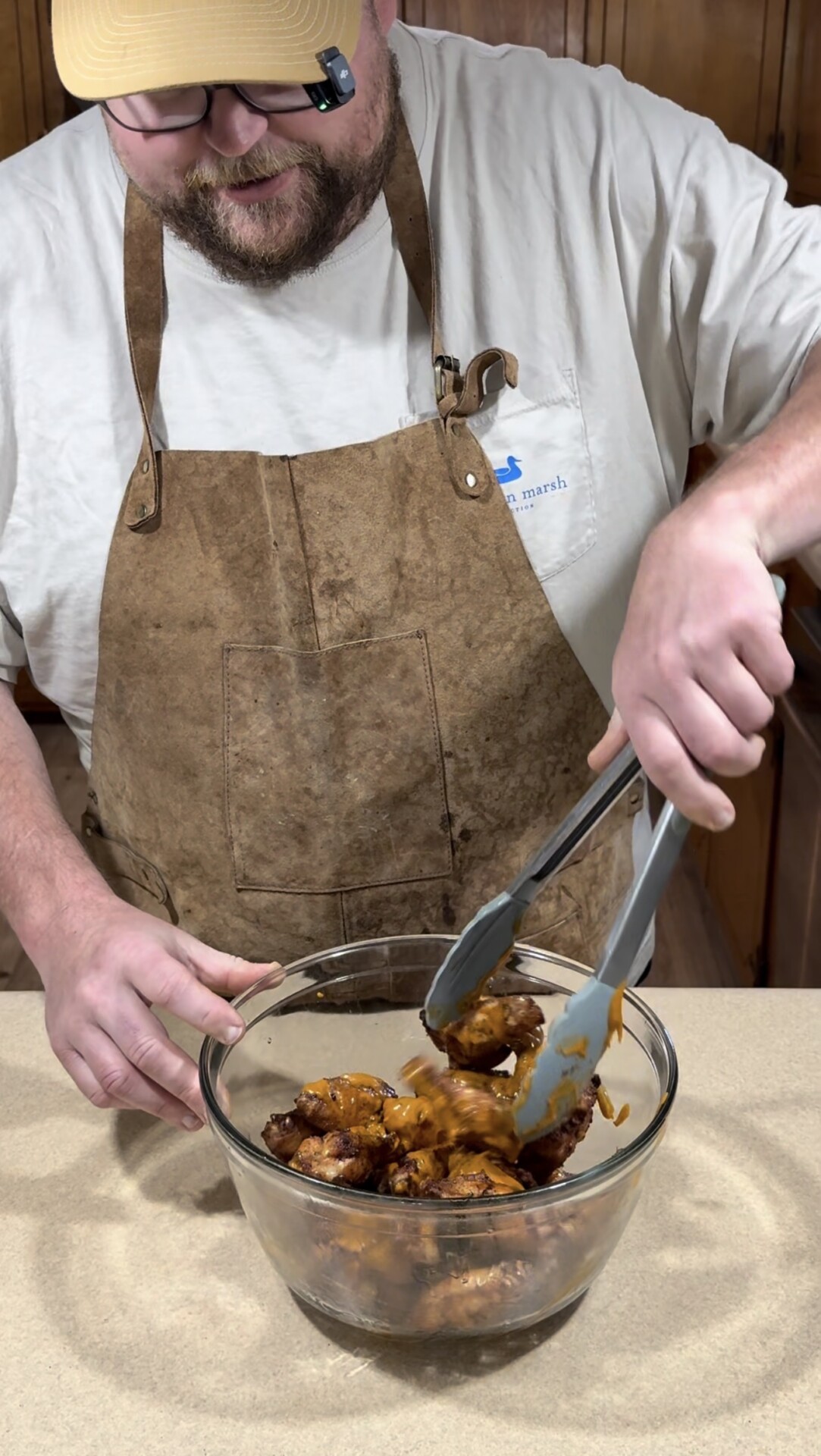 A person wearing a brown apron and white shirt uses tongs to mix sauced chicken wings in a glass bowl on a kitchen counter.