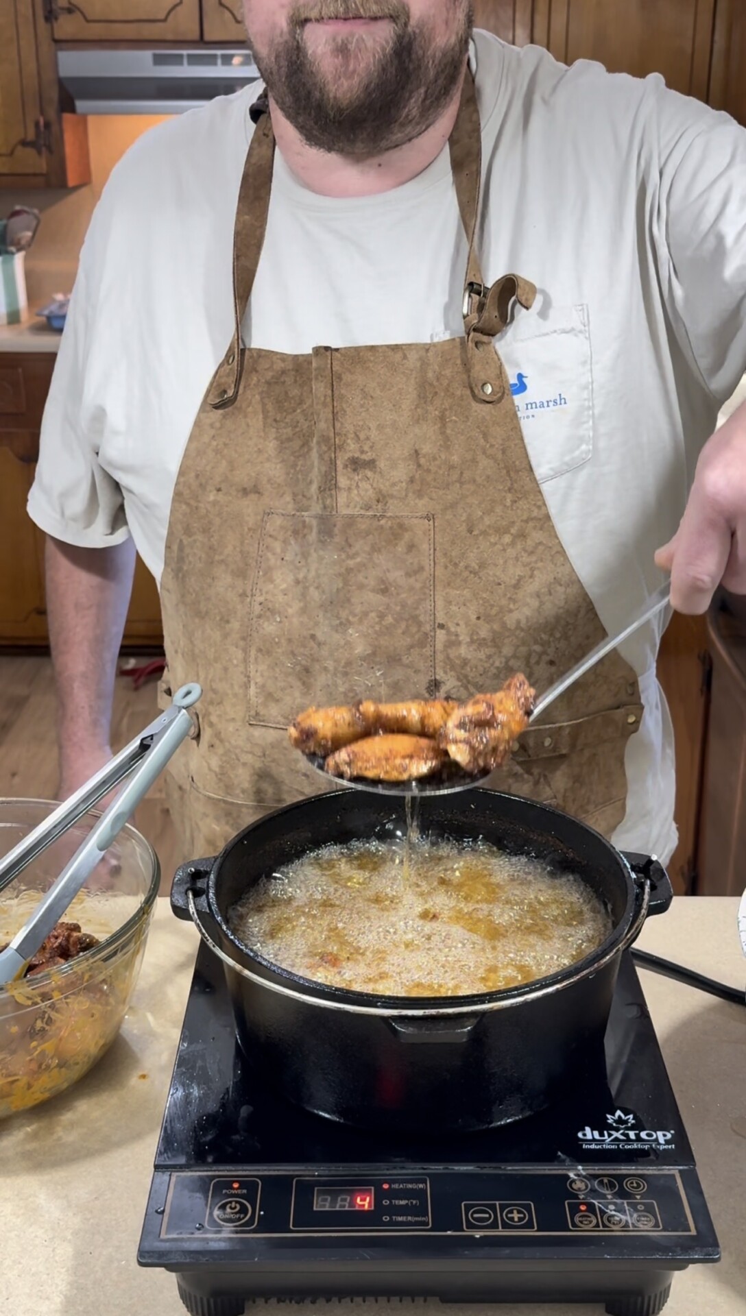 A person wearing a brown apron and white shirt is frying food in a black pot filled with oil. They hold up pieces of fried food with a slotted spoon, while tongs and a bowl of more food are nearby on the counter.