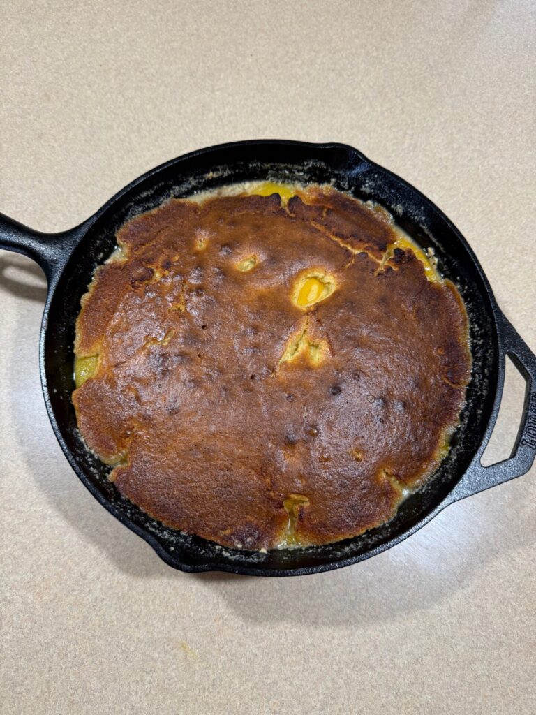 A golden-brown cornbread with slightly uneven coloring is baked in a black cast iron skillet, resting on a light-colored countertop.