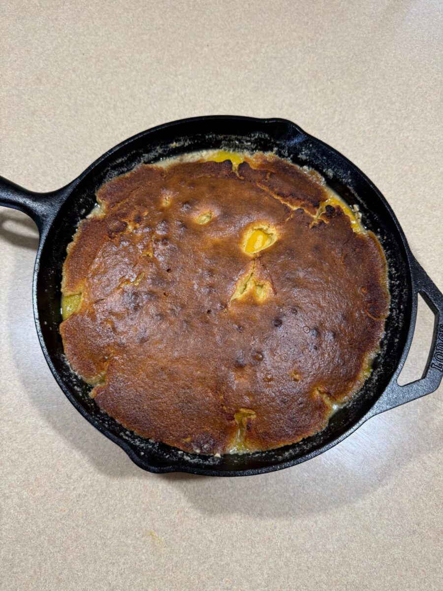 A golden-brown cornbread with slightly uneven coloring is baked in a black cast iron skillet, resting on a light-colored countertop.