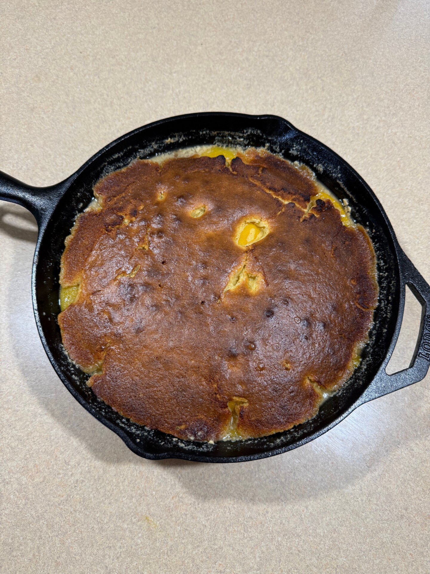 A golden-brown cornbread with slightly uneven coloring is baked in a black cast iron skillet, resting on a light-colored countertop.