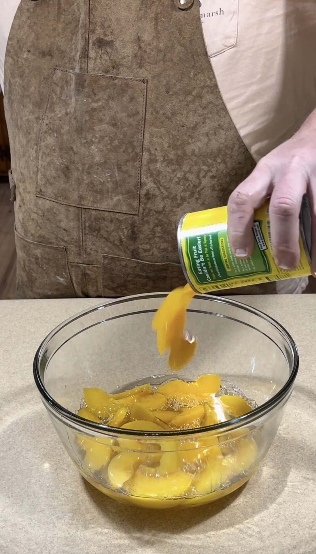 A person wearing a brown apron pours canned sliced peaches and syrup from a yellow can into a clear glass mixing bowl on a kitchen counter.