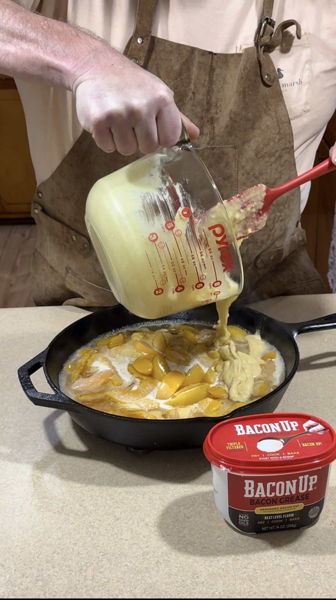 A person wearing a brown apron pours batter from a glass measuring cup into a skillet filled with sliced peaches. A container labeled BaconUp Bacon Grease sits on the countertop nearby.