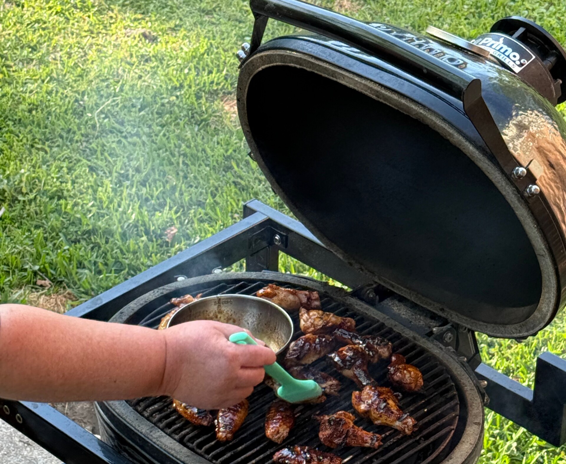 A person uses a green brush to apply sauce to chicken grilling on an outdoor barbecue, with smoke rising and grass visible in the background.