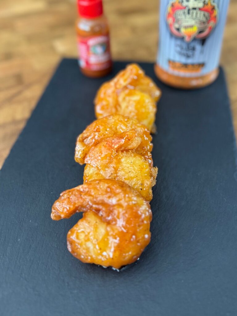Four glazed shrimp are arranged in a row on a black slate plate, with a hot sauce bottle and a seasoning container blurred in the background on a wooden surface.