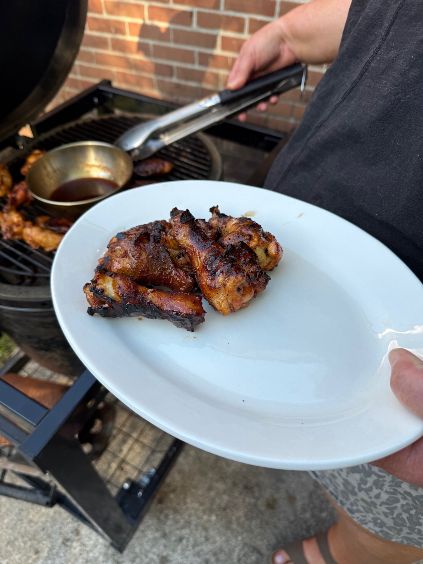 A person holds a white plate with grilled chicken pieces near a barbecue grill outdoors. A pan with sauce and more chicken are on the grill, and a brick wall is in the background.