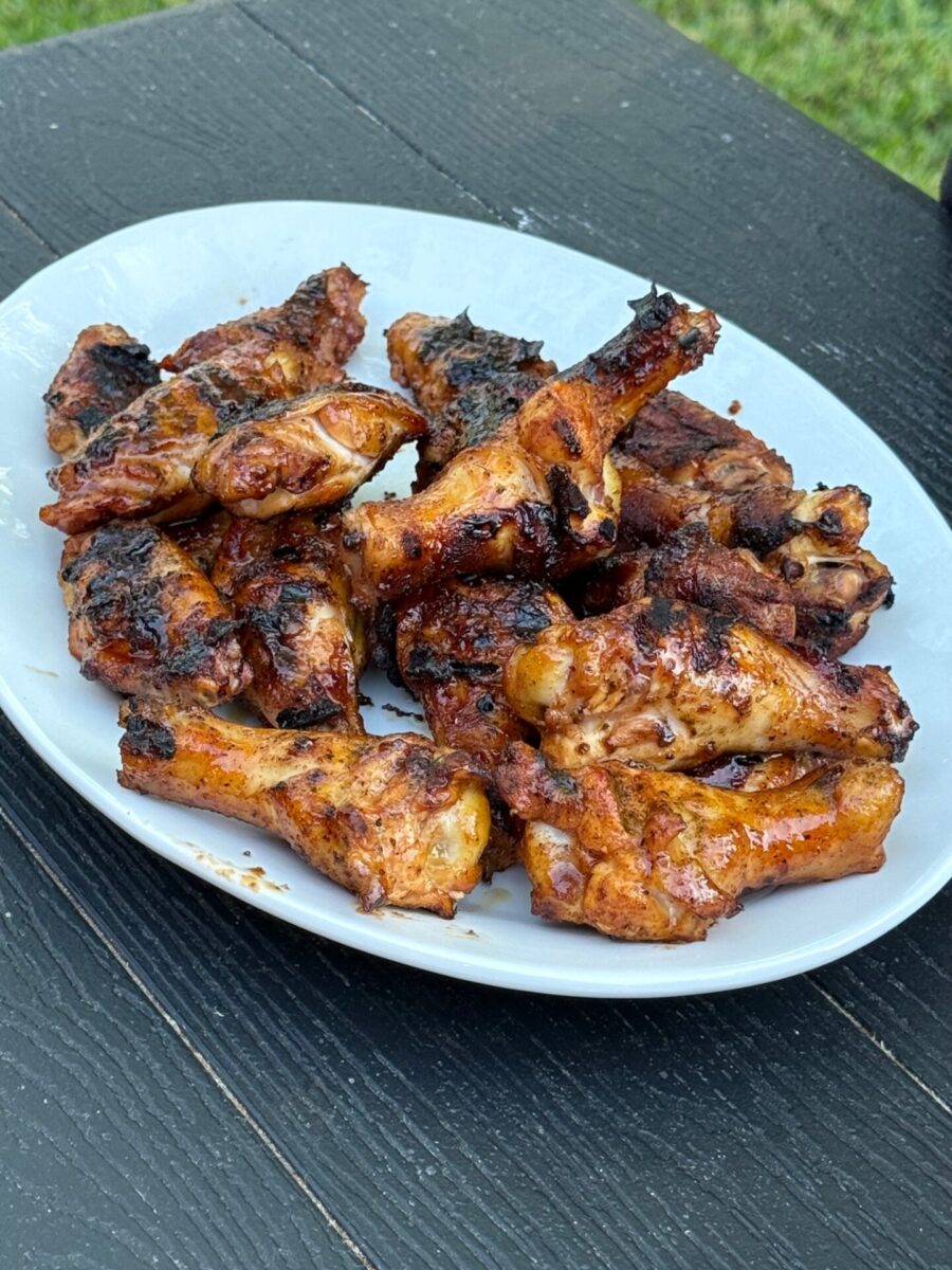 A white plate filled with grilled chicken wings sits on a dark wooden table outdoors, with some grass visible in the background. The wings are golden brown with charred, crispy edges.