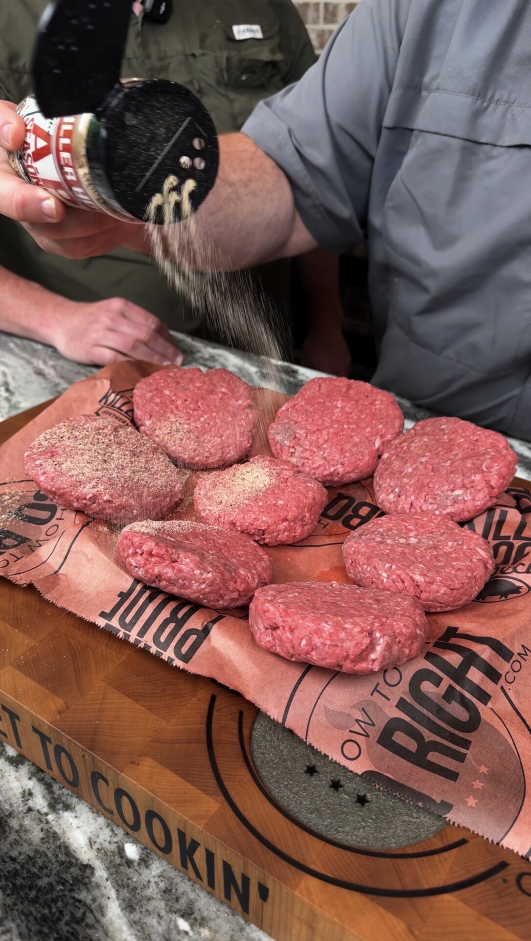 A person sprinkles seasoning from a shaker onto several raw ground beef patties arranged on butcher paper atop a wooden cutting board. Another person stands nearby, watching the process.