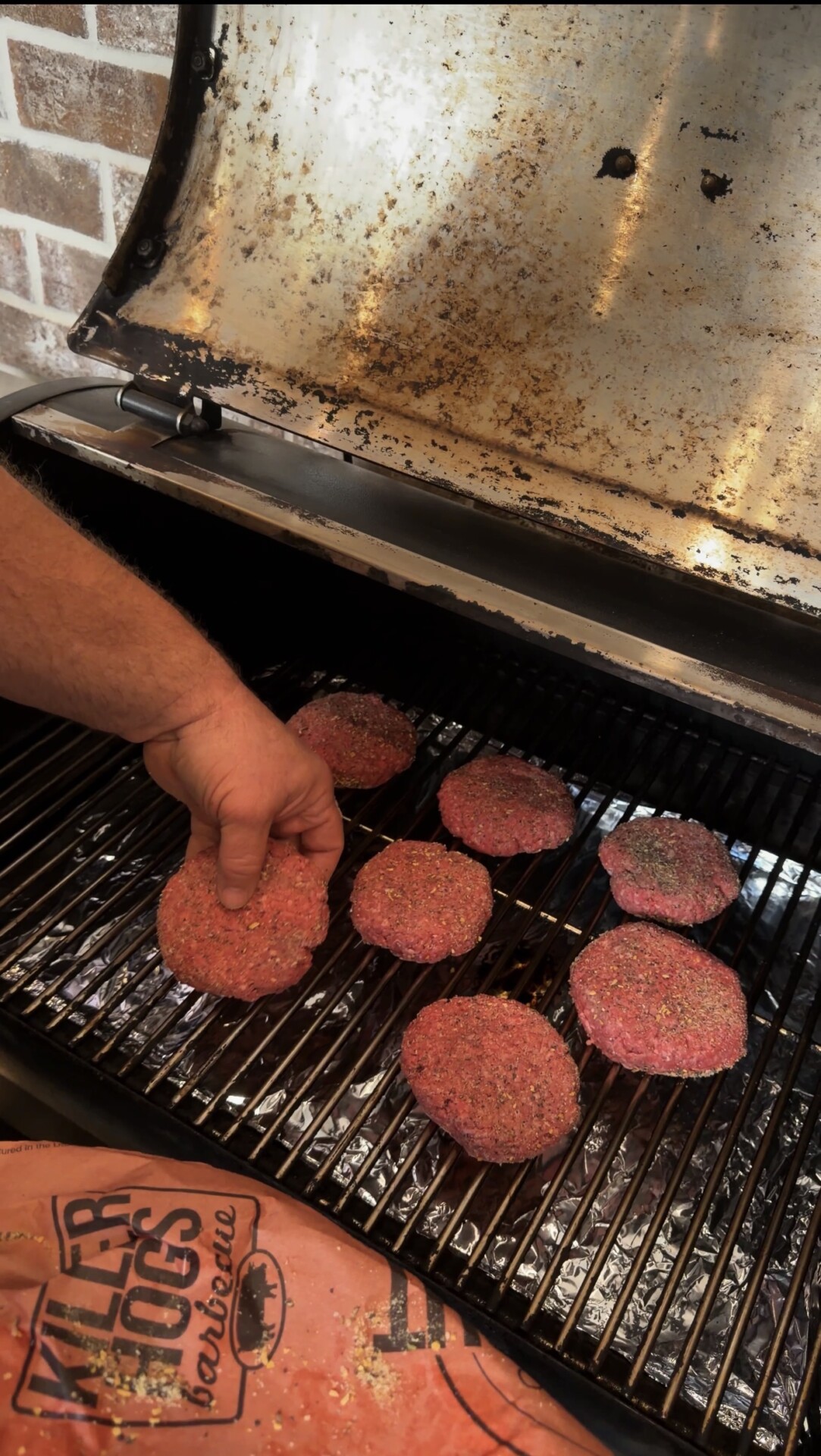 A person places seasoned raw hamburger patties on the grill of a barbecue smoker, preparing them for cooking. Several patties are already on the grill, and the smoker lid is open.