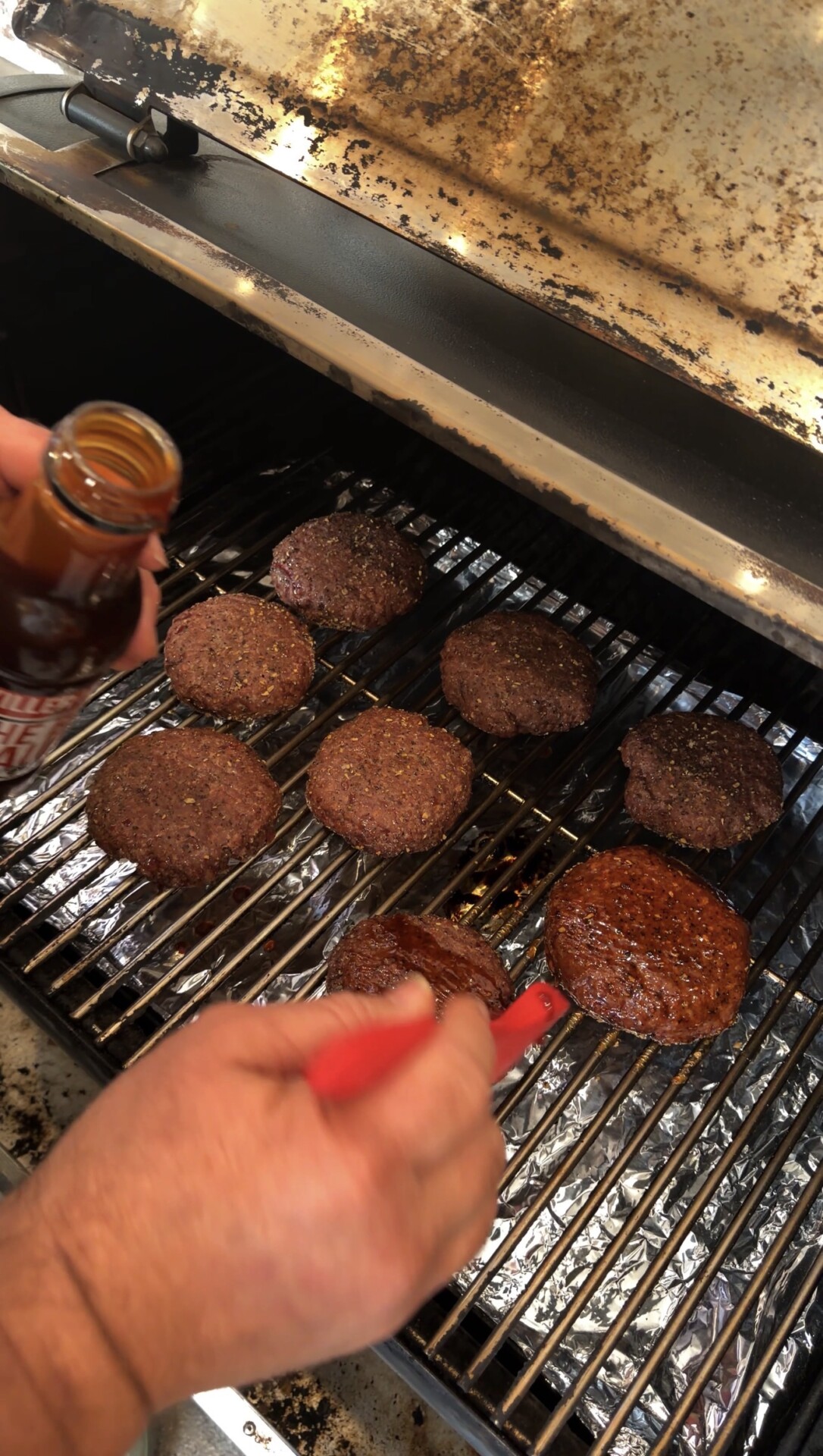 A person brushes barbecue sauce onto burger patties while they cook on a grill, holding a bottle of sauce in one hand and a basting brush in the other.