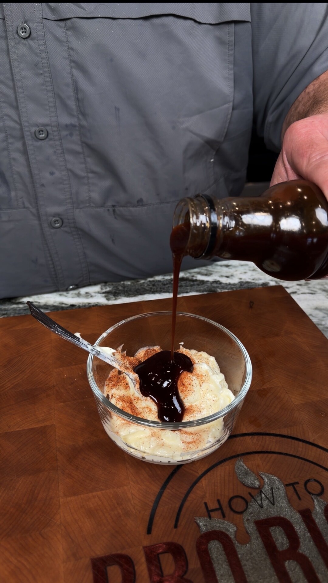 A person pours dark barbecue sauce from a bottle onto a bowl of coleslaw sprinkled with paprika, with a spoon in the bowl, on a wooden cutting board.