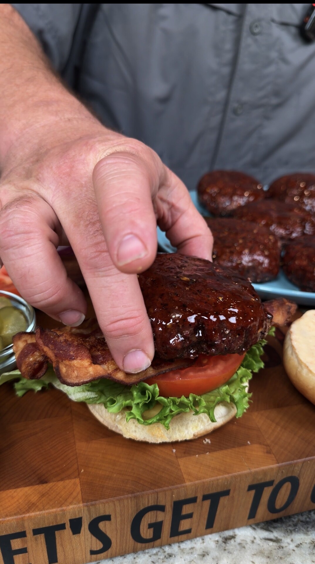A close-up of a person assembling a burger with lettuce, tomato, bacon, and a glazed burger patty on a bun, set on a wooden board. The persons hand is placing the patty on the burger.