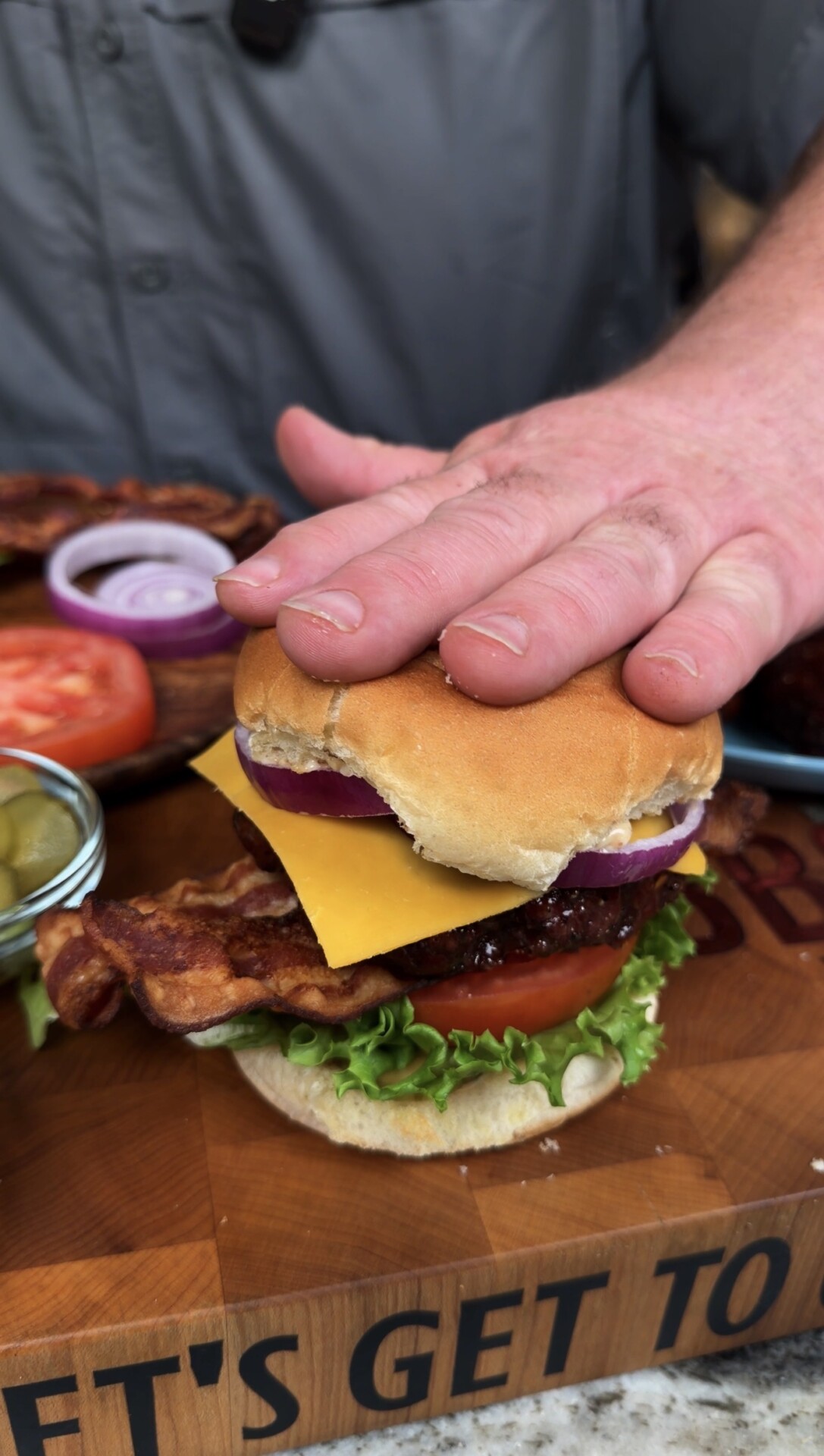 A hand presses down on a stacked bacon cheeseburger with lettuce, tomato, onion, and cheddar on a bun. Sliced tomatoes, onions, and pickles are visible on the wooden board nearby.