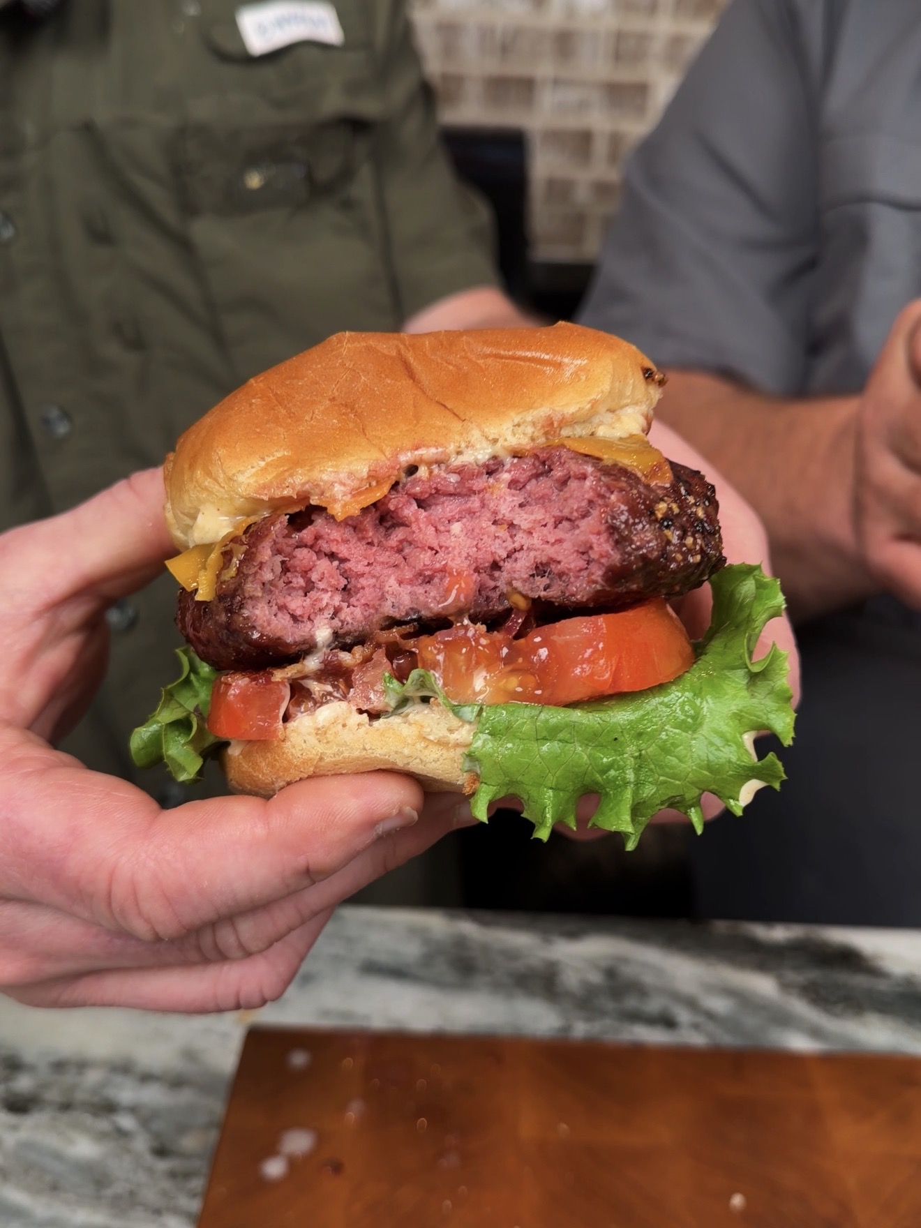A person holds a hamburger with a thick, juicy beef patty cooked medium-rare, topped with lettuce, tomato, melted cheese, and a soft bun. Another person stands beside them, partially visible.