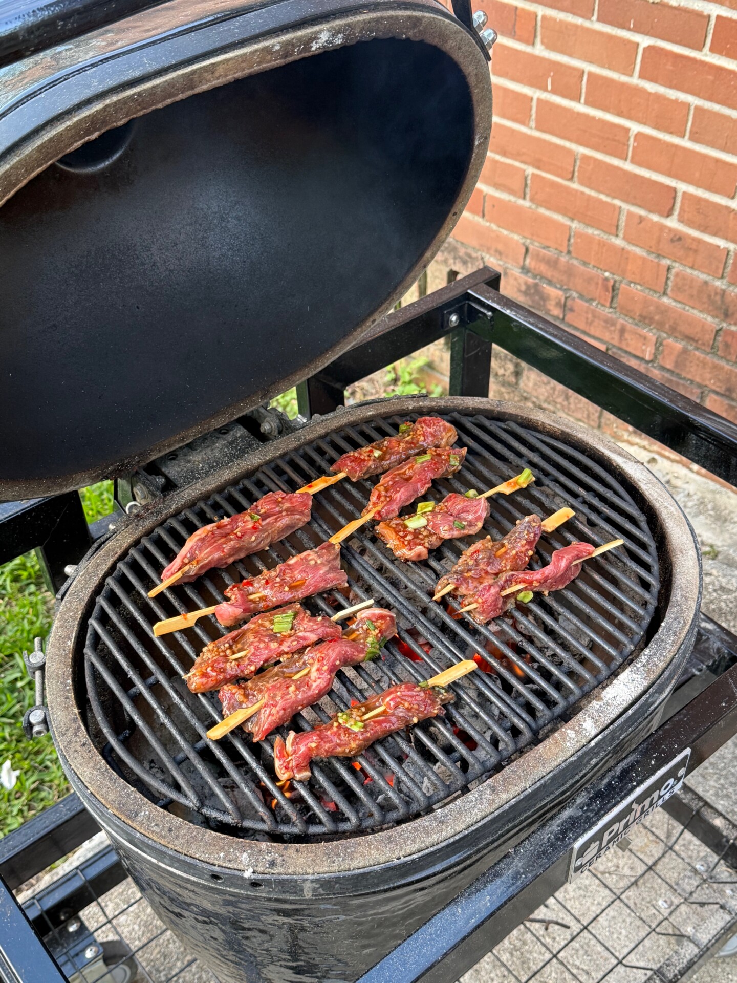 Raw marinated beef skewers are placed on the grill grate of a black ceramic charcoal grill, with the lid open. The grill sits outdoors near a red brick wall and some green grass.
