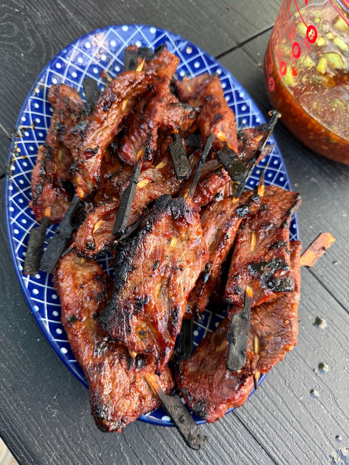 A blue patterned plate piled with grilled skewered meat, charred and glazed, sitting on a dark wooden table. A glass bowl of reddish-brown dipping sauce with sliced green onions is visible to the side.