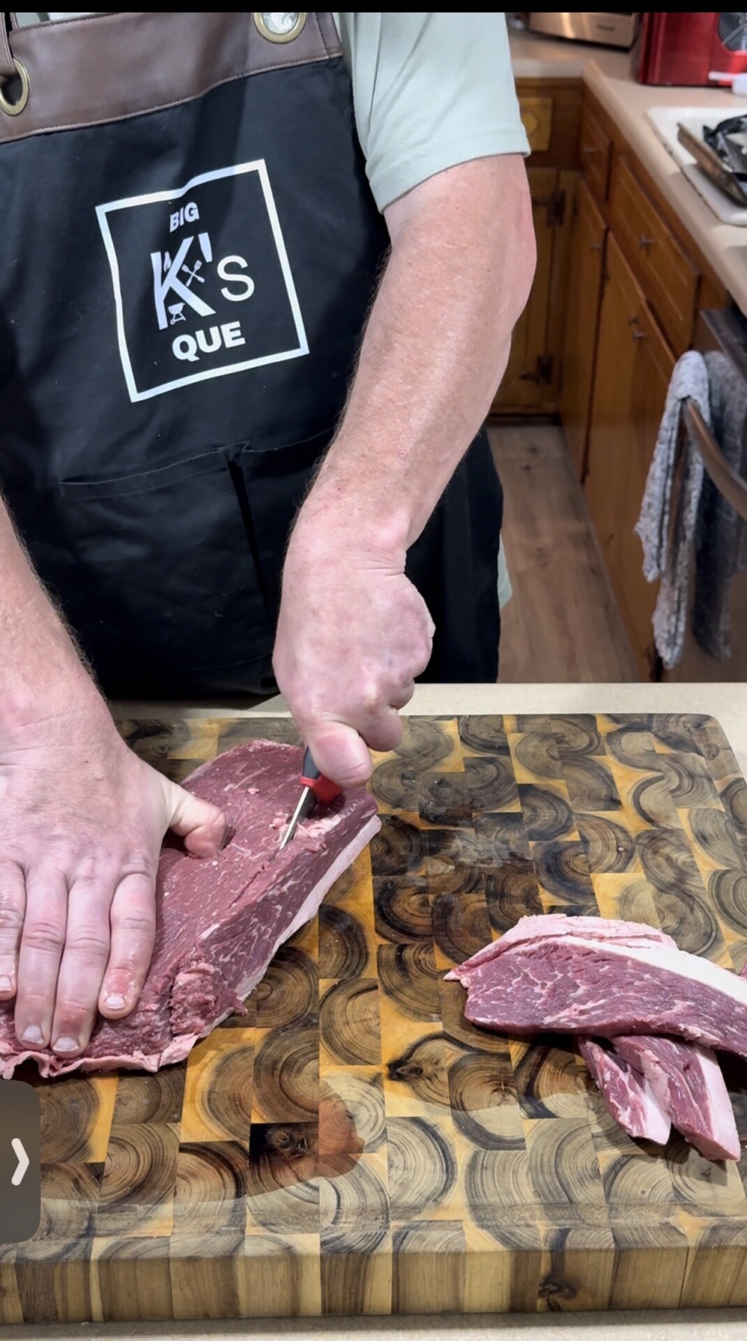 A person wearing a Big Ks Que apron slices a raw steak on a wooden cutting board, with several pieces of meat already cut off to the side.
