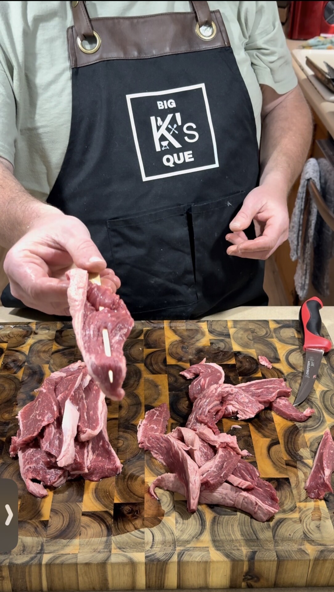A person wearing a Big K’s Que apron holds up a slice of raw beef over a wooden cutting board with more sliced beef and a red-handled knife.