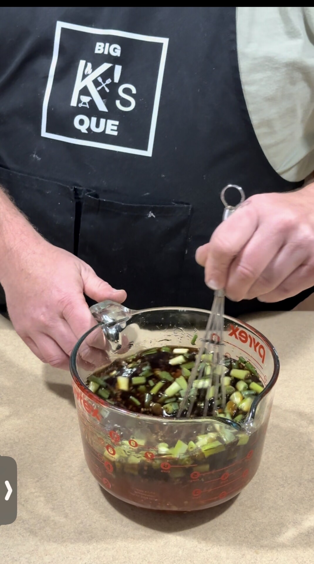 A person wearing a black apron labeled BIG KS QUE is whisking a mixture of chopped green onions and dark sauce in a glass Pyrex measuring cup.