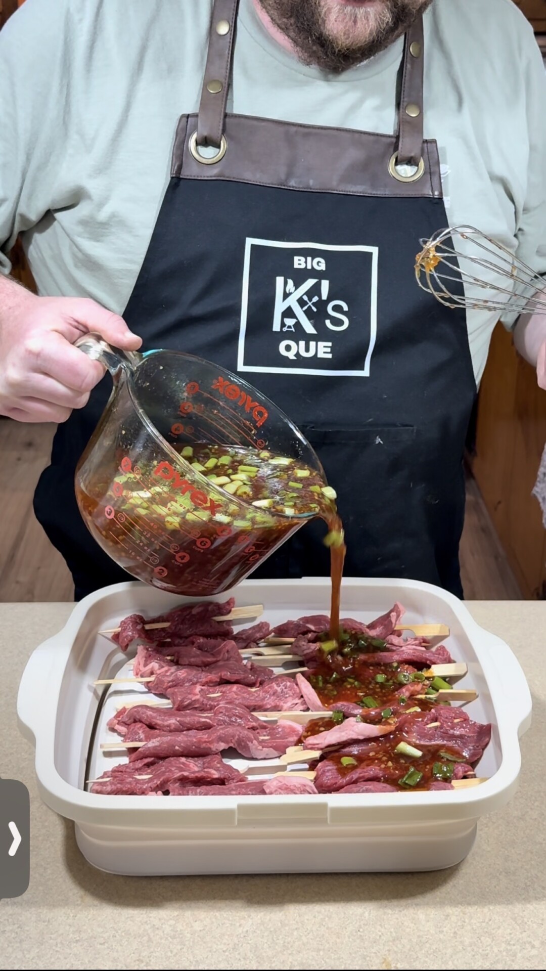 A person wearing a BIG KS QUE apron pours a marinade with chopped vegetables from a measuring jug over raw skewered meat in a baking dish on a kitchen counter.