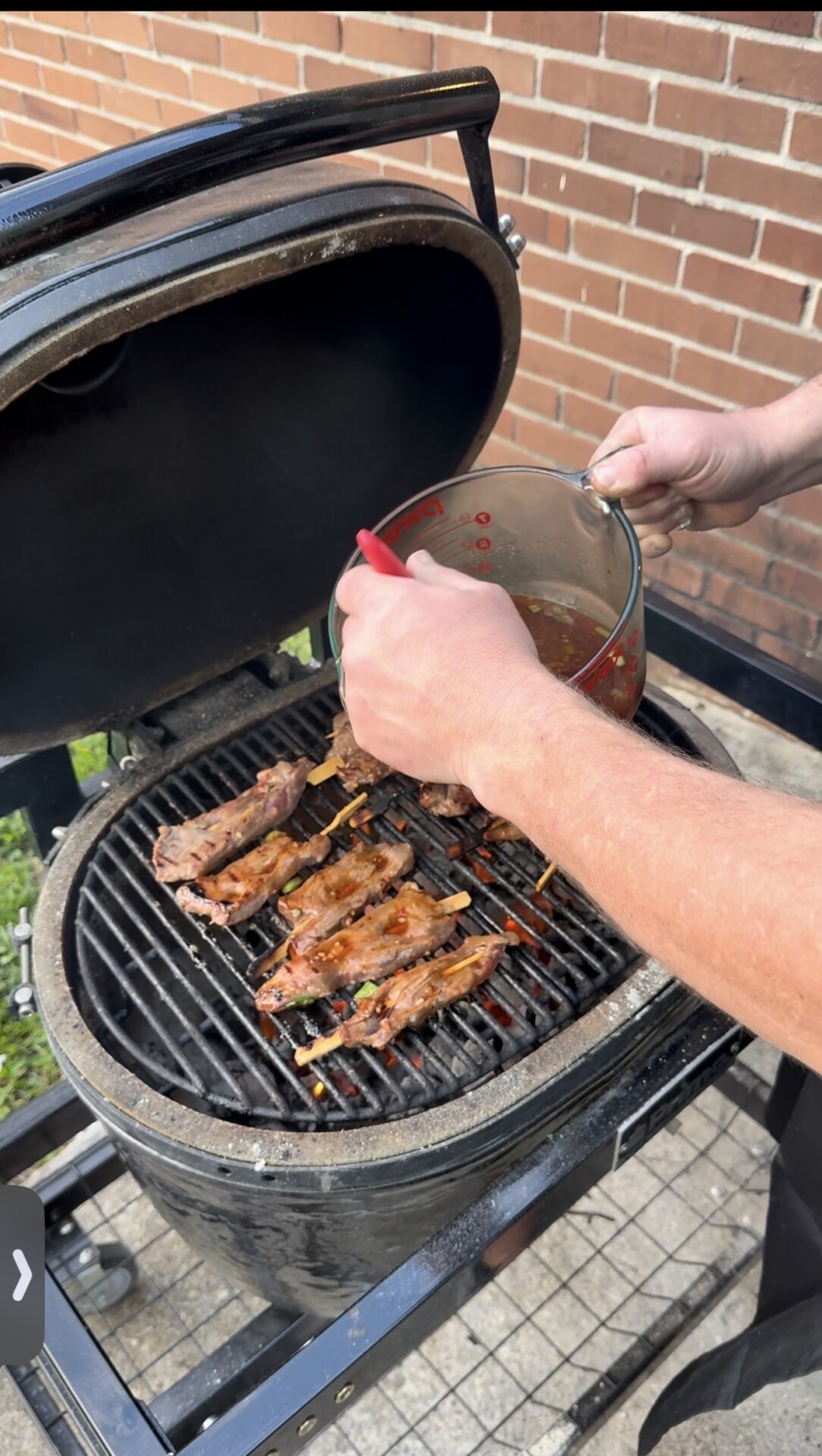 A person pours marinade from a glass measuring cup onto skewered meat cooking on a round charcoal grill, set outdoors next to a brick wall.