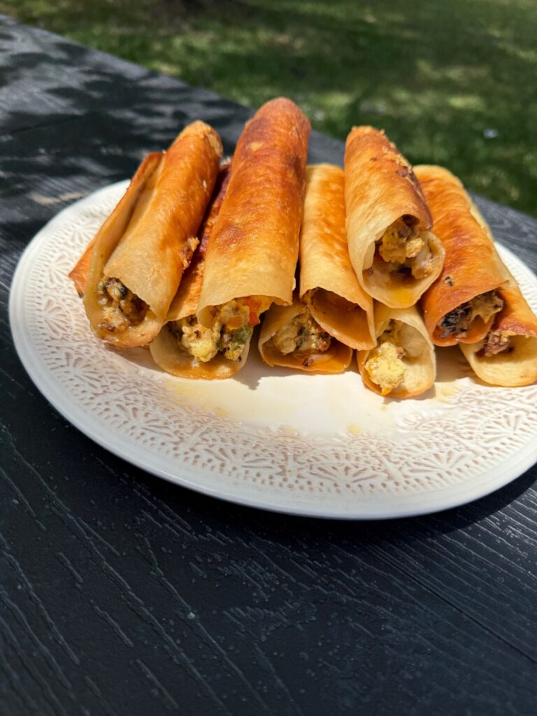 A plate with seven golden-brown, crispy rolled taquitos filled with a cheesy and possibly meat or veggie mixture, placed on a dark outdoor table with grass visible in the background.