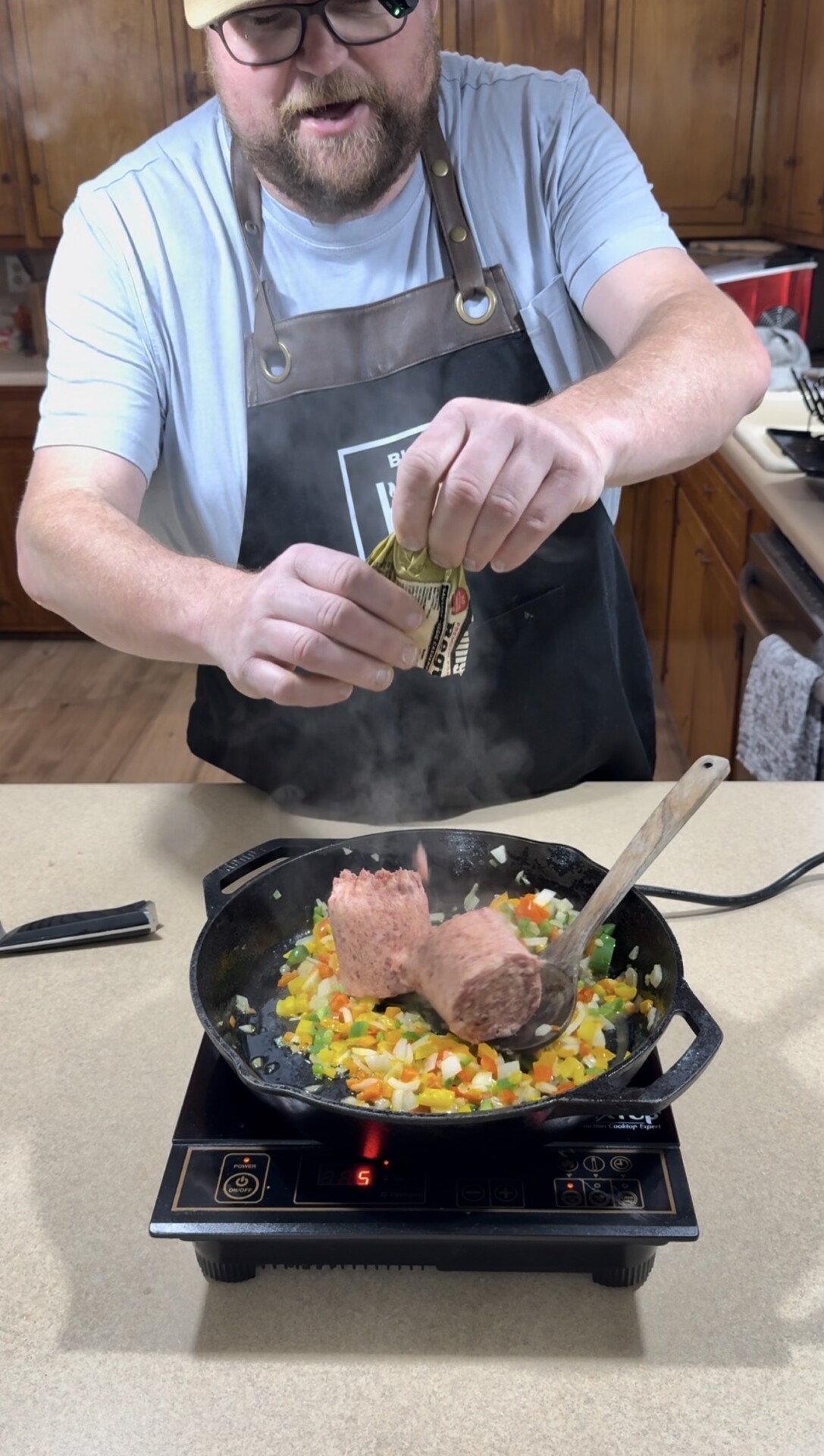 A person wearing glasses and an apron is cooking in a kitchen, adding ground meat and seasoning to a skillet filled with chopped vegetables on an electric stove. Steam rises from the hot pan.