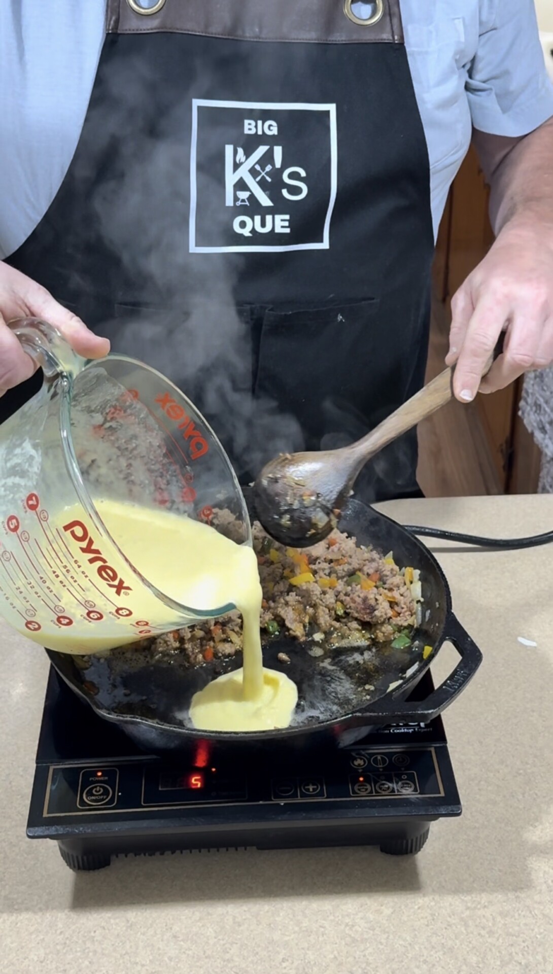 A person wearing a “Big K’s Que” apron pours beaten eggs from a glass measuring cup into a hot skillet with cooked meat and vegetables, stirring with a wooden spoon over an induction cooktop. Steam rises from the pan.