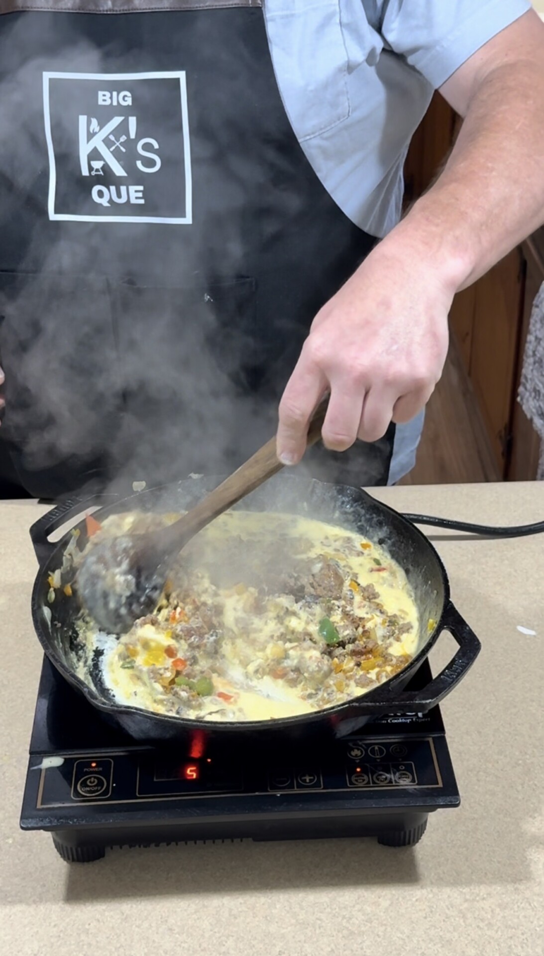 A person wearing a BIG KS QUE apron stirs a creamy, colorful mixture of vegetables and meat in a cast iron skillet on an electric stovetop, with steam rising from the pan.