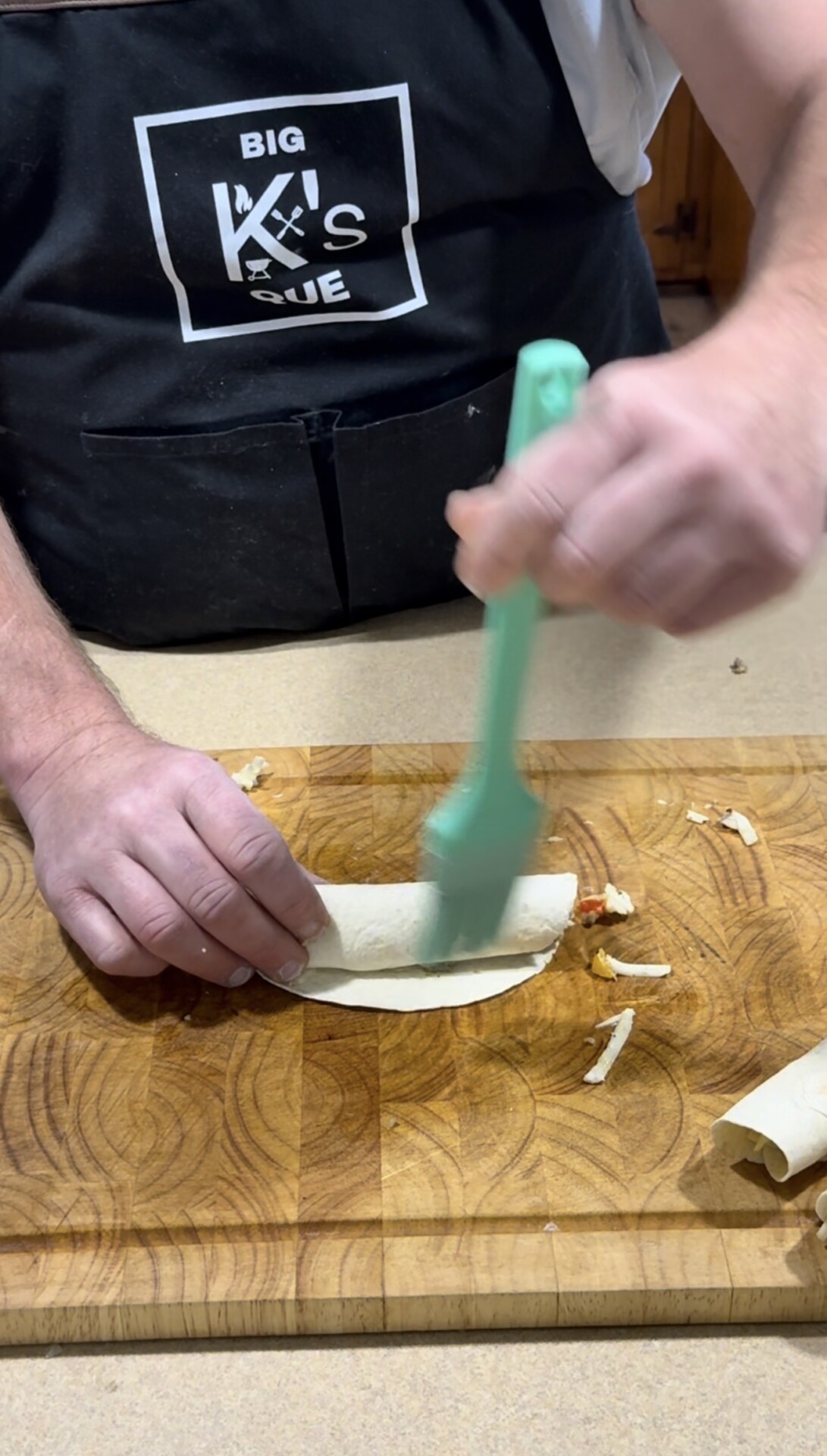 A person wearing a black apron is brushing a rolled pastry with a green silicone brush on a wooden cutting board. Pastry trimmings are scattered nearby.