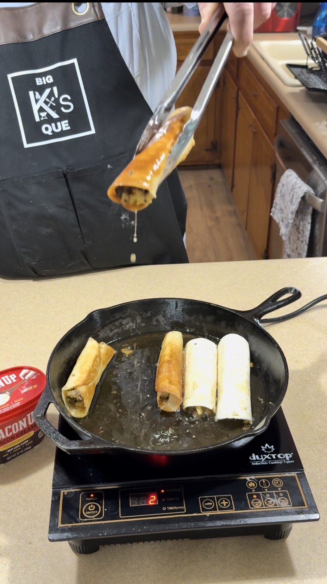 A person wearing a Big Ks Que apron uses tongs to remove a fried taquito from a cast iron skillet with three more taquitos cooking on an induction cooktop. An open can of bacon grease sits nearby.