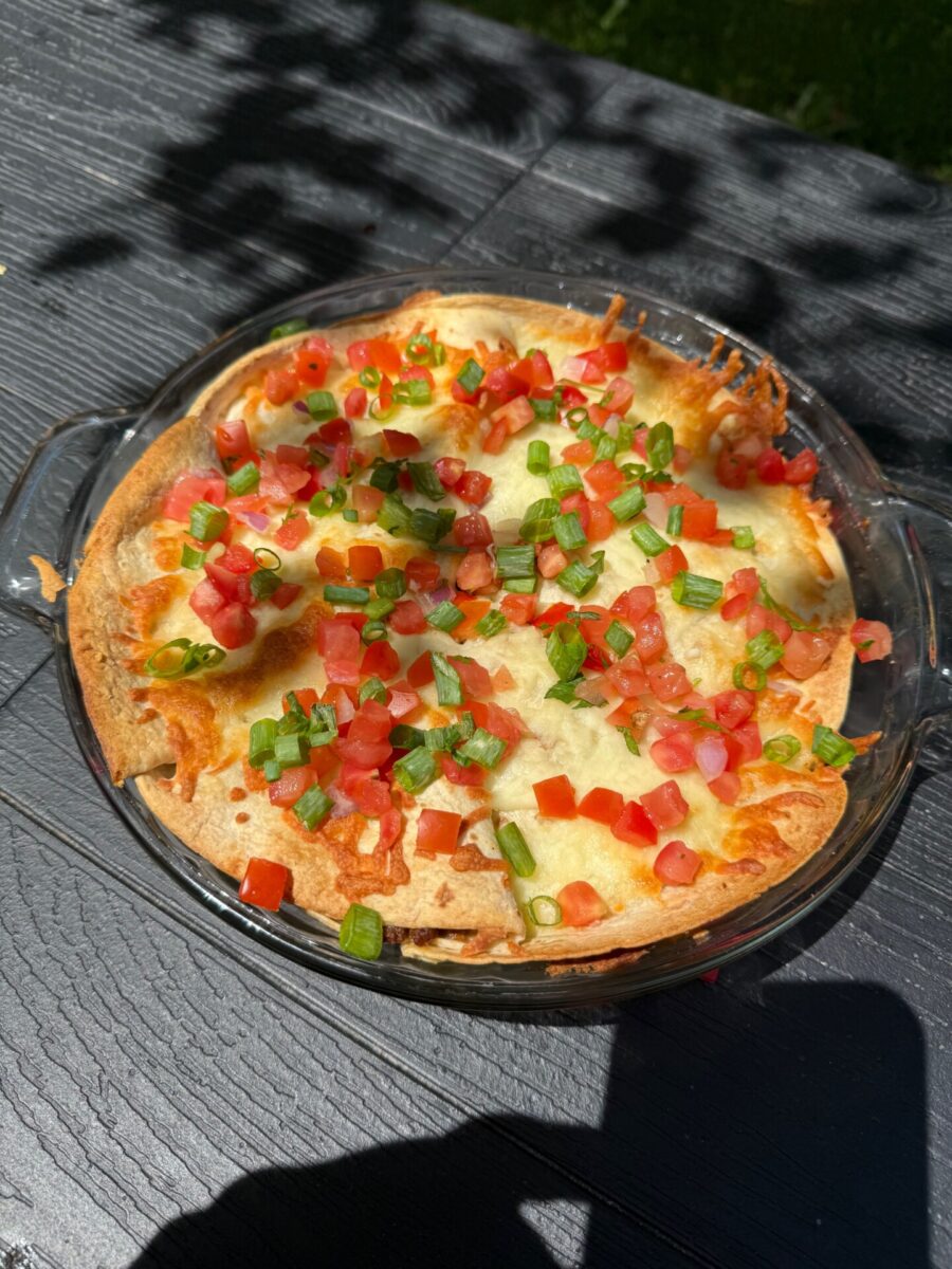 A round glass dish filled with a baked cheesy casserole topped with diced tomatoes and chopped green onions, sitting on a dark wooden outdoor table in sunlight.