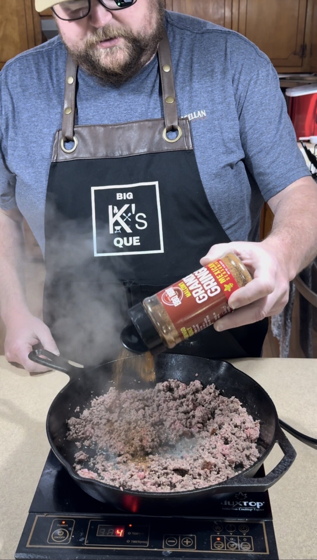 A person wearing a “Big K’s Que” apron is seasoning browned ground meat in a cast iron skillet with a bottle of Grillin’ Gravy seasoning. Steam rises from the pan on a stovetop.