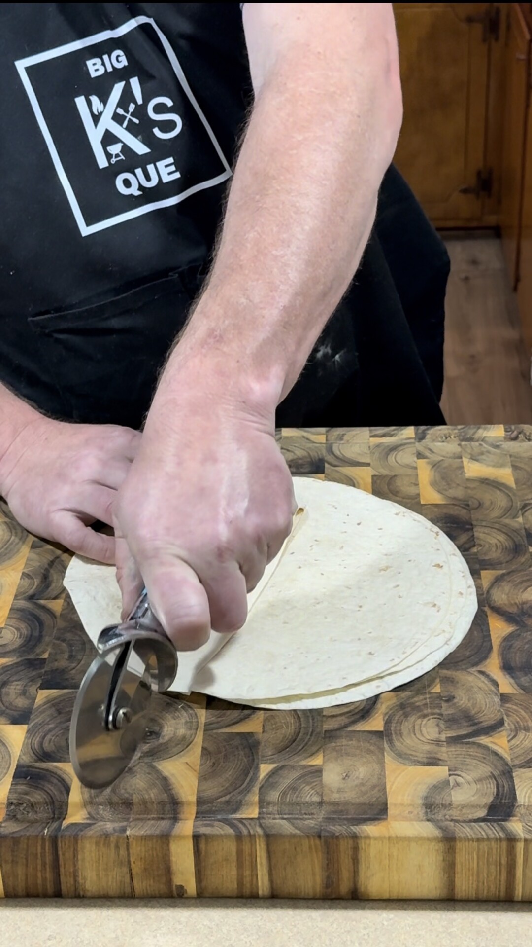 A person wearing a Big Ks Que apron uses a pizza cutter to slice a flour tortilla on a wooden cutting board.