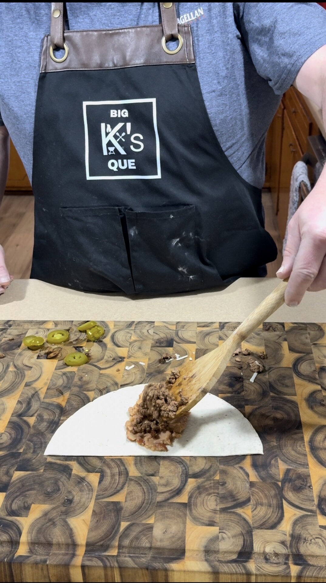 A person in a black “Big K’s Que” apron uses a wooden spoon to add a meat mixture onto a tortilla on a patterned cutting board, with sliced jalapeños nearby.
