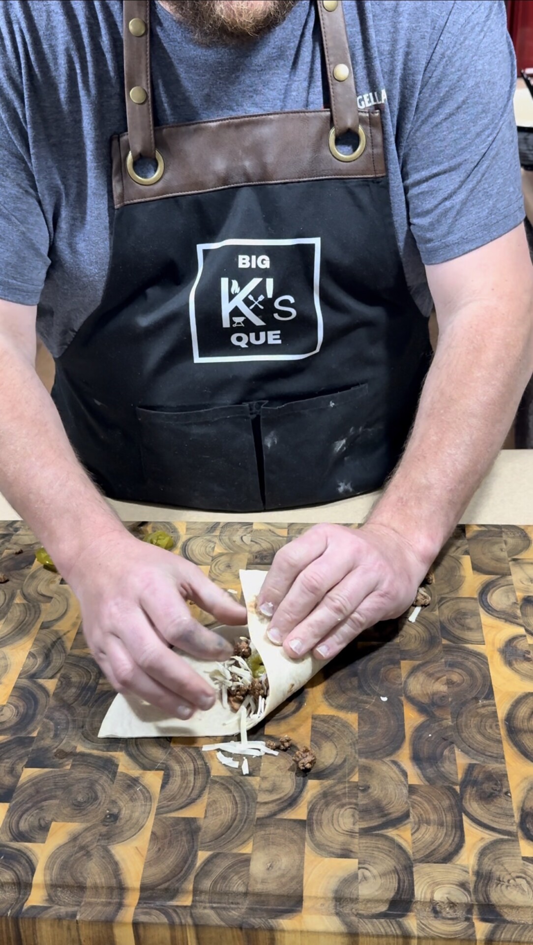 A person wearing a Big K’s Que apron folds a tortilla wrap with shredded meat and cheese on a patterned wooden cutting board. Only their hands and torso are visible.