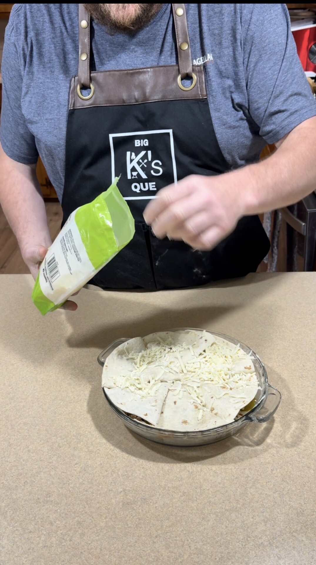 A person wearing a black apron labeled BIG KS QUE is sprinkling shredded cheese from a bag onto tortillas in a glass baking dish on a kitchen counter.