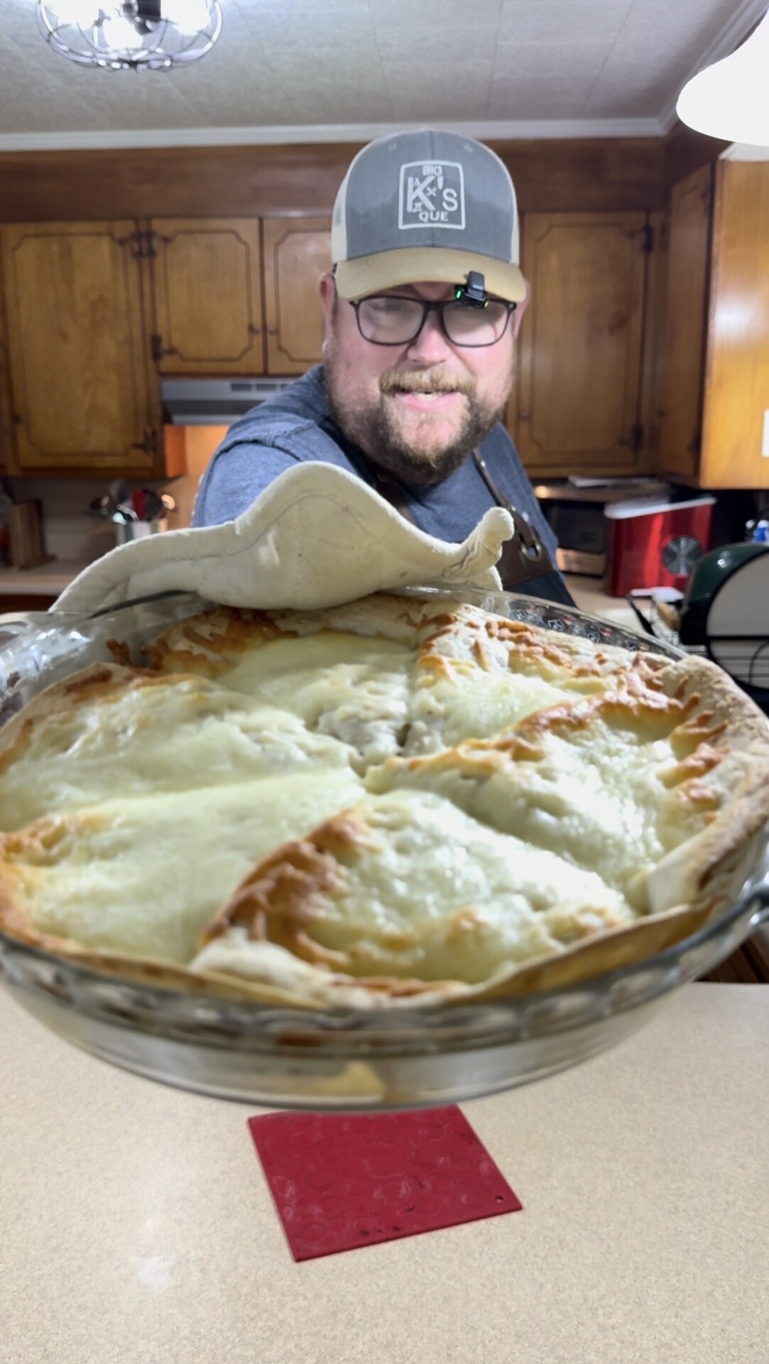 A smiling man in a kitchen holds a freshly baked pie in a glass dish towards the camera, with a white cloth draped over his hand and wooden cabinets in the background.
