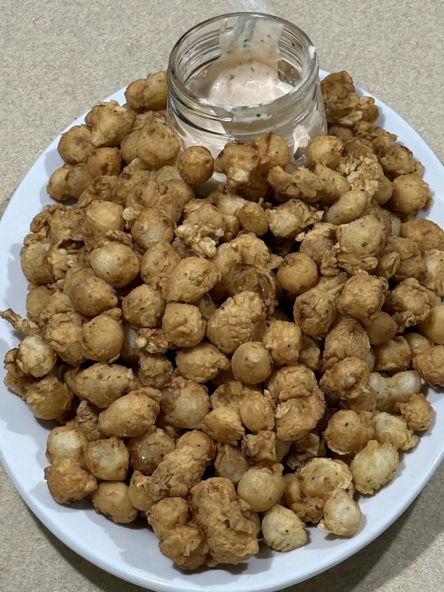 A white plate filled with a pile of small, golden-brown fried bites, served with a partially empty glass jar of creamy dipping sauce in the center. The plate rests on a beige countertop.