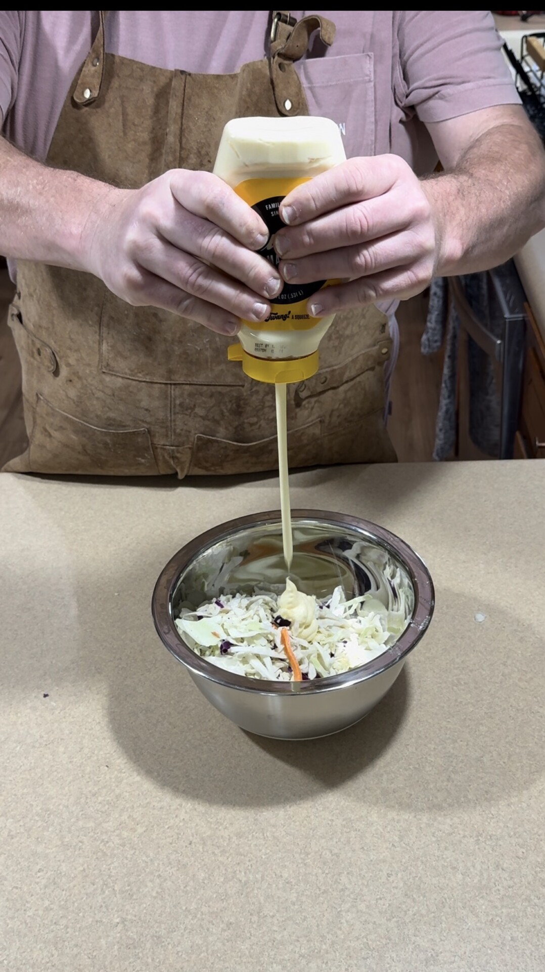 A person in a brown apron squeezes yellow mustard from a bottle onto a bowl of shredded cabbage and carrots on a kitchen counter.