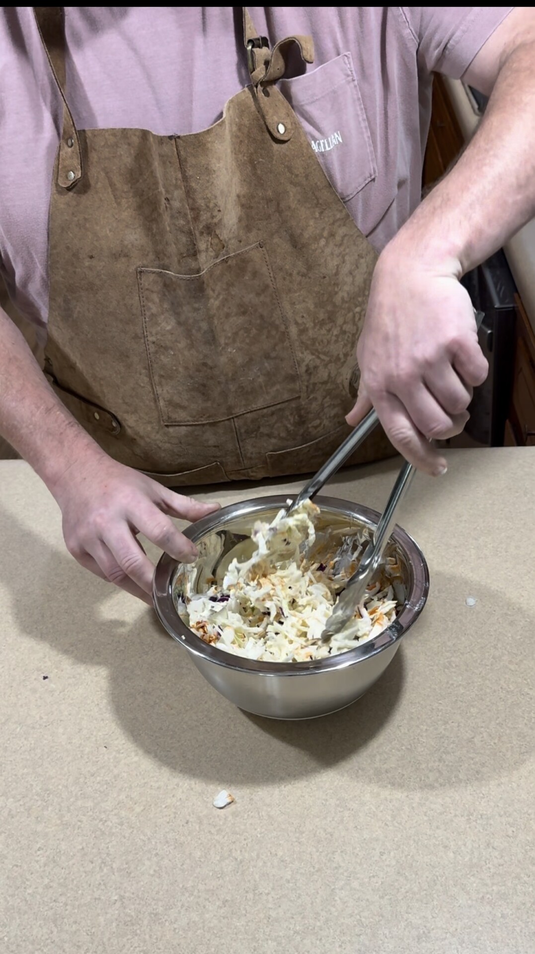 A person wearing a brown apron is mixing shredded coleslaw with tongs in a stainless steel bowl on a beige countertop.