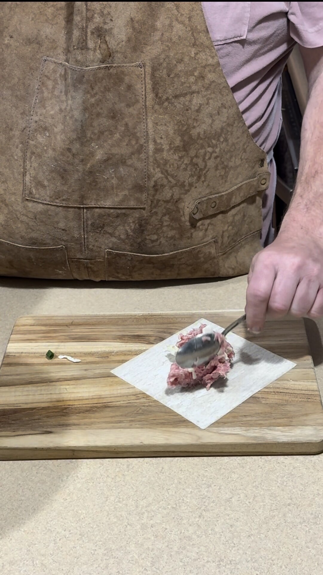 A person wearing a brown apron is spooning ground meat onto a square dumpling wrapper on a wooden cutting board, preparing to make dumplings.