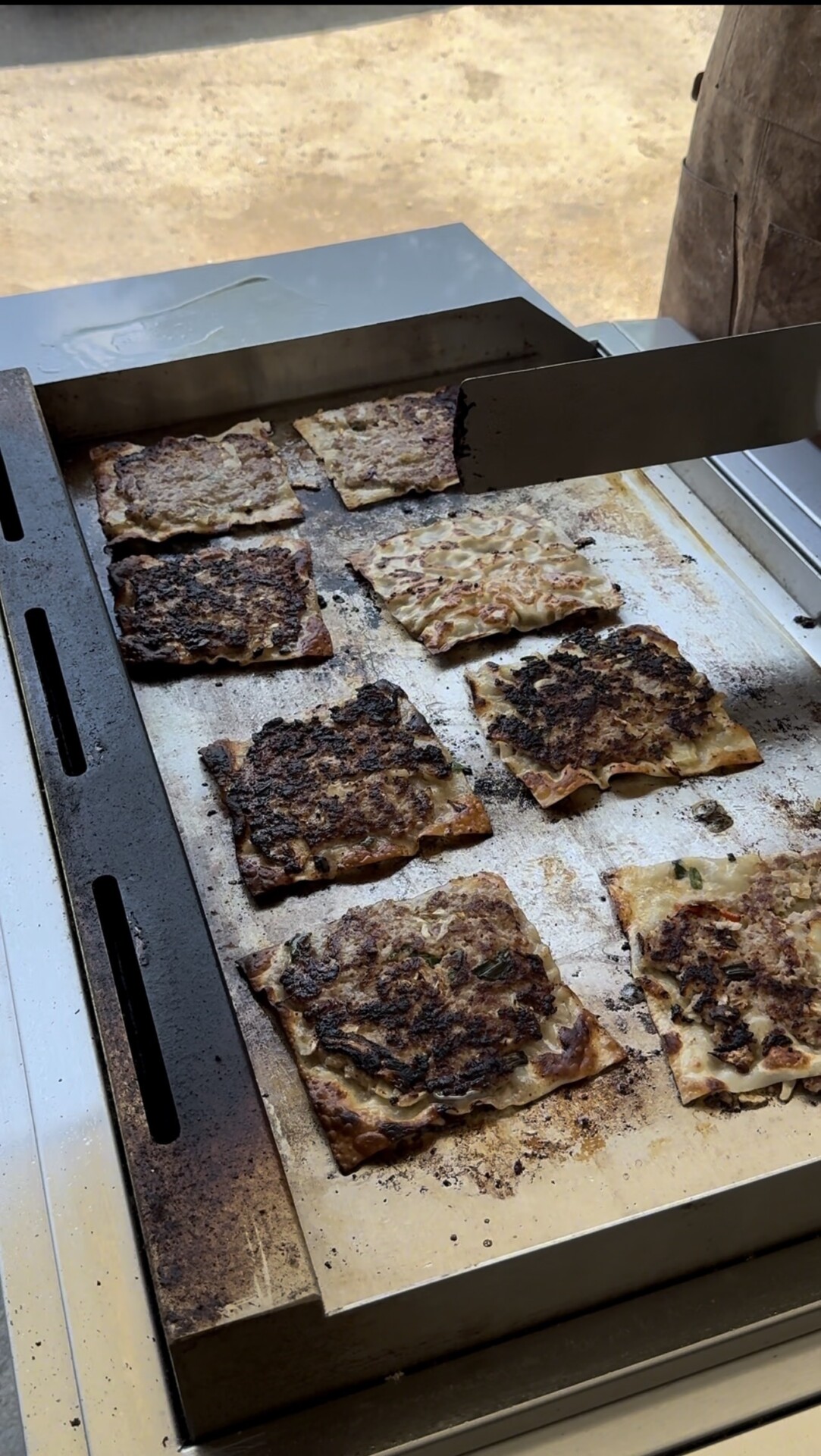 Rectangular pieces of flatbread or savory pancakes are being cooked on a large metal griddle, some with visible browning and toppings like vegetables or meat. A metal spatula rests on the griddle’s edge.