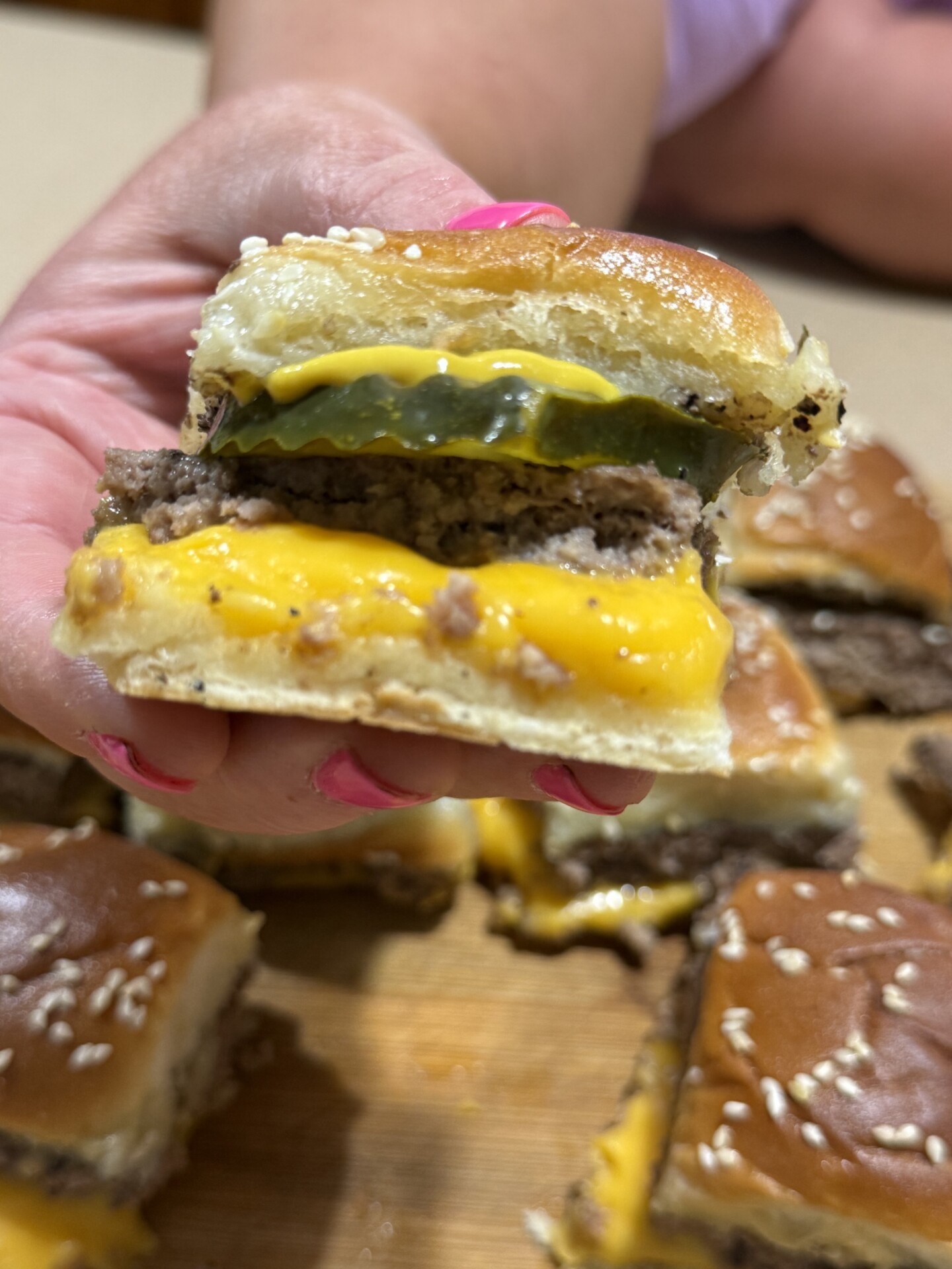 A hand with pink-painted nails holds a small cheeseburger slider with melted cheese, pickles, and a sesame seed bun. More sliders are visible on a wooden surface in the background.