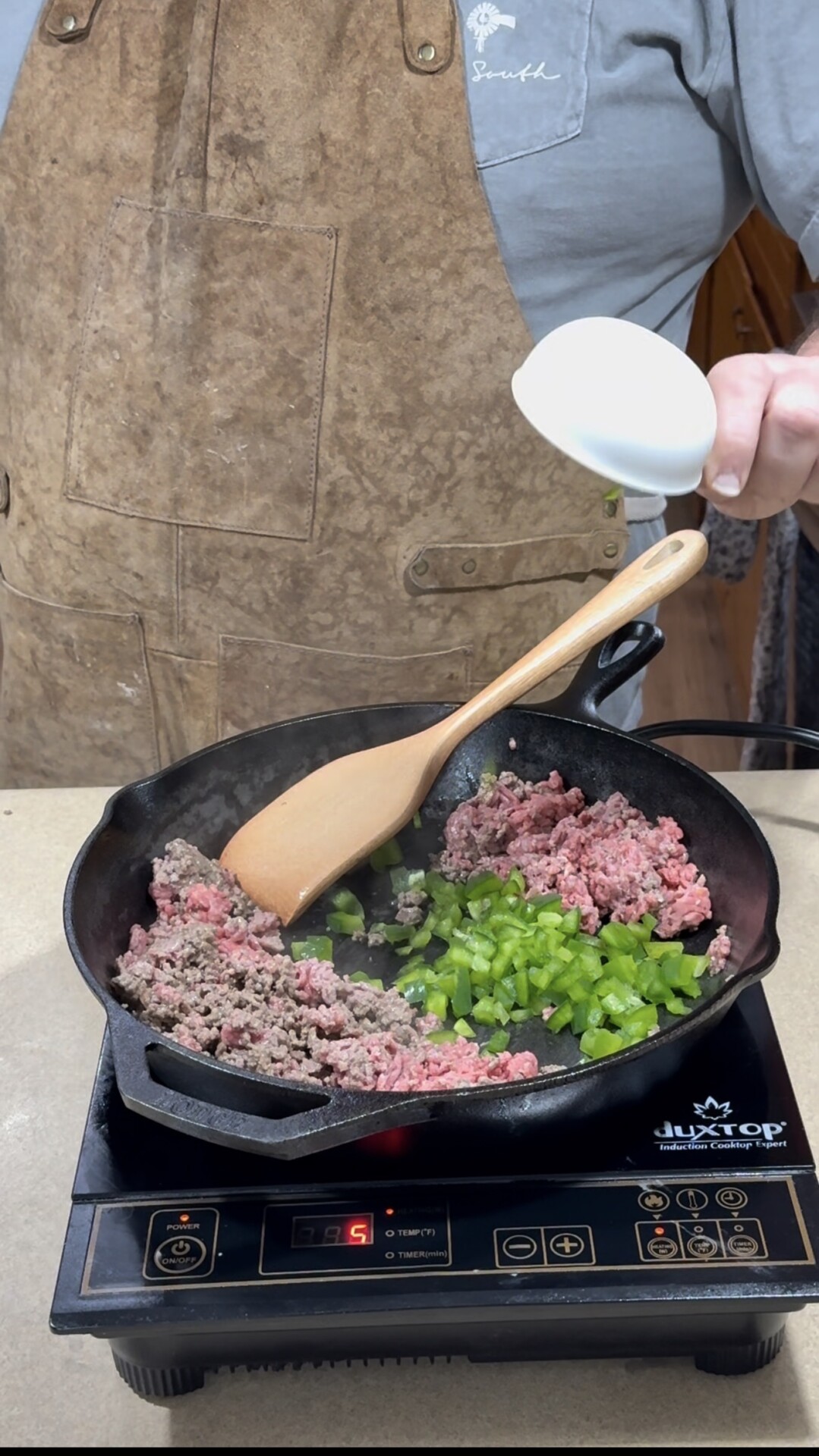 A person in a brown apron cooks ground beef and chopped green bell peppers in a cast iron skillet on an induction cooktop, stirring with a wooden spoon and holding a small white bowl above the pan.