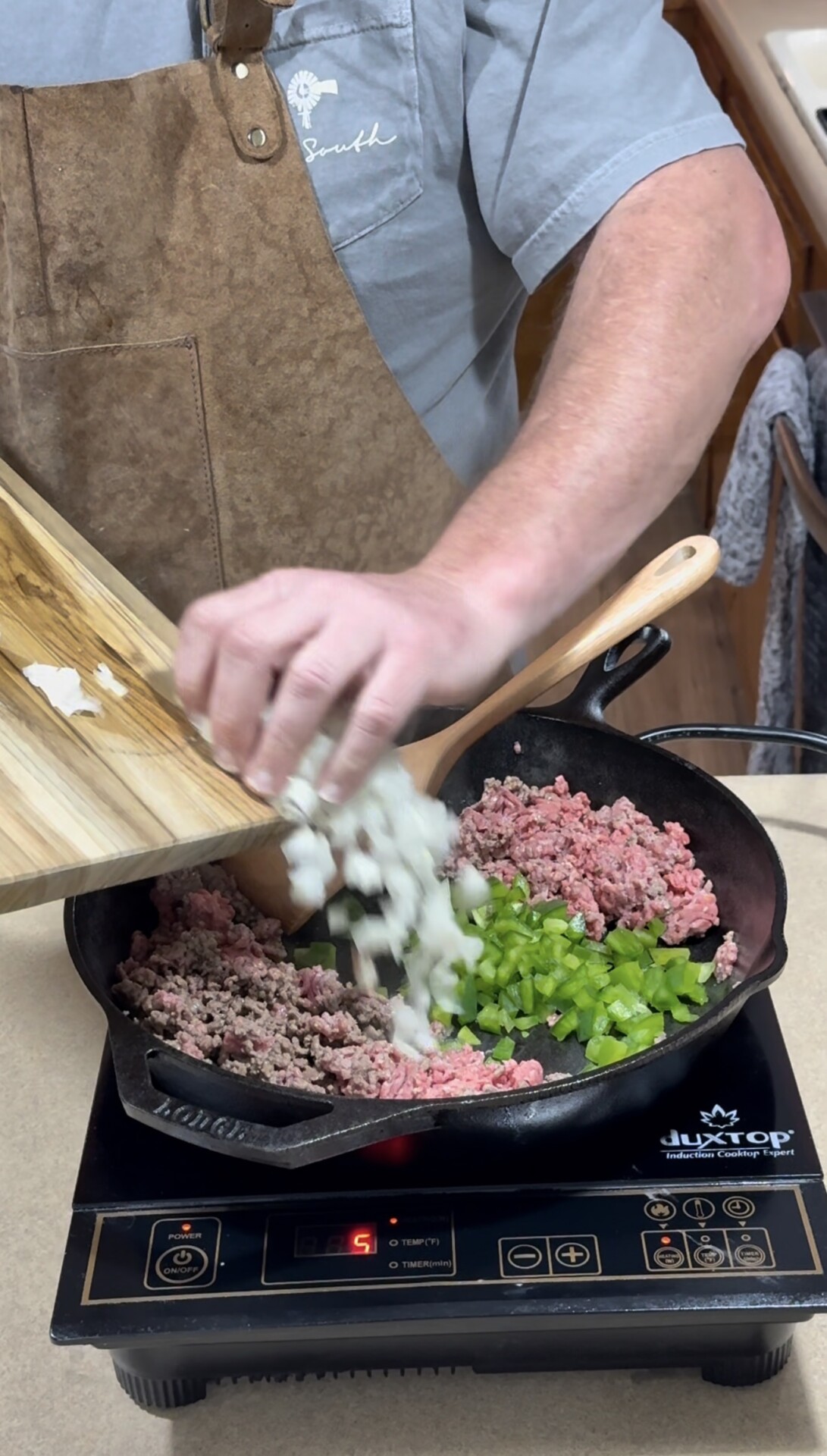 A person wearing a brown apron adds chopped onions from a cutting board into a cast iron skillet with ground meat and diced green bell peppers on an induction cooktop.